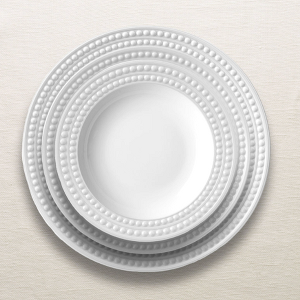 Four white ceramic plates from L'Objet's Perlèe White Dinnerware collection, each featuring a beaded rim, are neatly stacked on a light, textured surface.