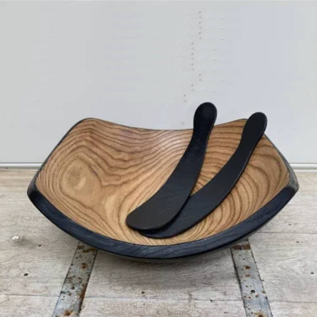 The Peterman Ebonized Oak Salad Tosser Set rests inside a square wooden bowl with natural grain and black rim, creating a striking dining accent on a rustic surface against a plain backdrop.
