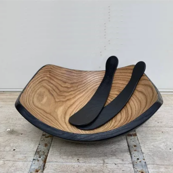 The Peterman Ebonized Oak Salad Tosser Set rests inside a square wooden bowl with natural grain and black rim, creating a striking dining accent on a rustic surface against a plain backdrop.