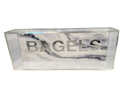 The Eye Candy Bagel Tray is a rectangular, food-safe acrylic tray featuring "BAGELS" in large silver letters on the front, set against a white and silver marble-effect acrylic background.
