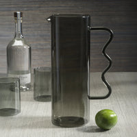 The Zodax Casa Smoke Pitcher, featuring a tall gray glass body and wavy handle, stands beside two glasses and a tequila bottle, with a lime in the foreground.