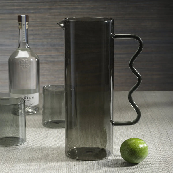 The Zodax Casa Smoke Pitcher, featuring a tall gray glass body and wavy handle, stands beside two glasses and a tequila bottle, with a lime in the foreground.