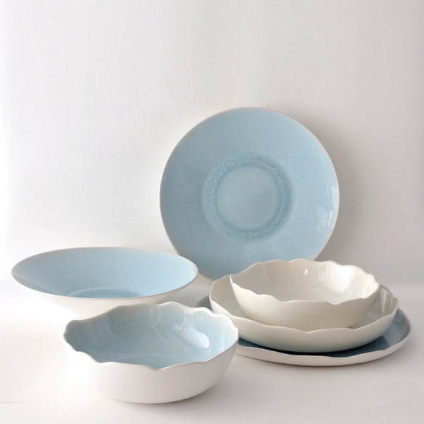 The Plume Ocean Blue dinnerware collection by Jars - Rosenthal features ceramic dishes and bowls in white and light blue, with smooth and wavy edges, highlighting artisanal craftsmanship against a plain white background.