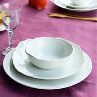 A place setting on a purple tablecloth features white scalloped ceramic pieces from the Plume White Dinnerware Pearl collection by Jars - Rosenthal, with a gold fork, a glass of lemon water, and an orange in the background.