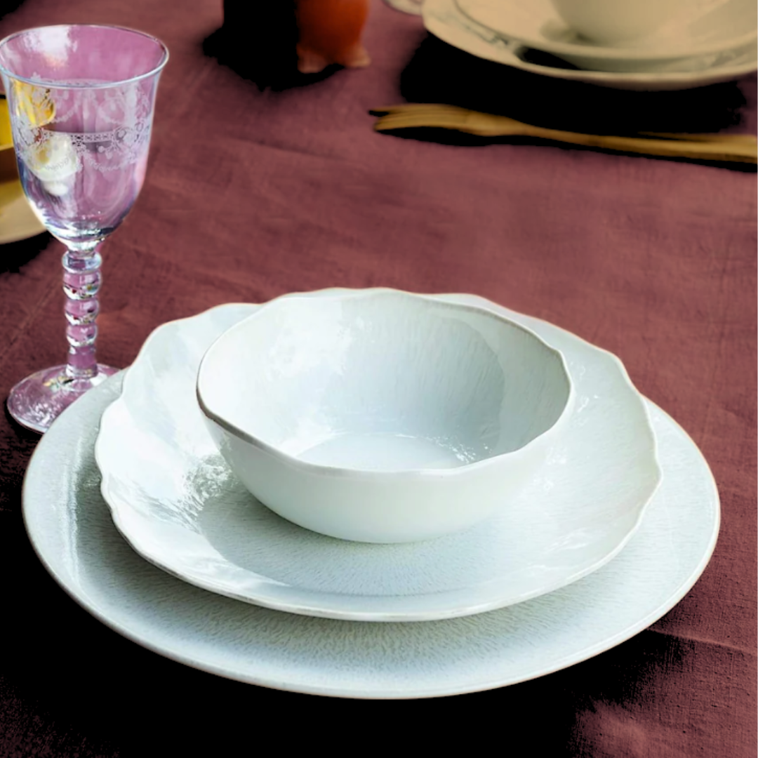 Plume White Dinnerware Pearl by Jars - Rosenthal, including a bowl, salad plate, and dinner plate, is stacked on a maroon tablecloth beside a gold fork and clear flower-etched wine glass. Another elegant set appears in the background.