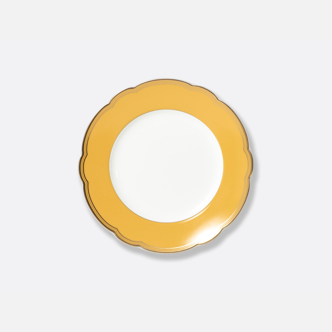 The Pompadour by Bernardaud is a round plate with a wide, mustard-yellow scalloped rim and white center, inspired by classic Limoges designs, shown against a plain light gray background.
