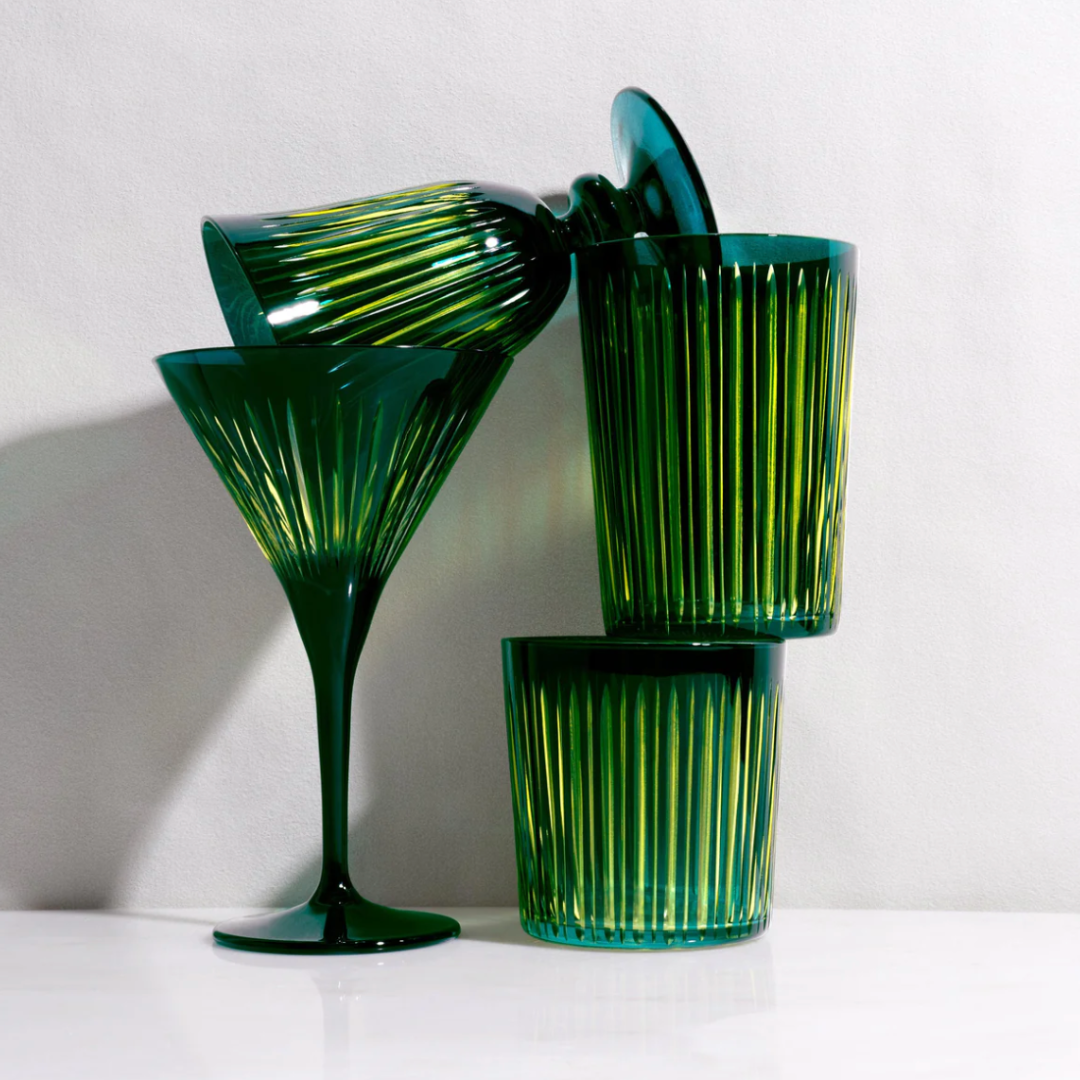 Four green-tinted, hand-cut Prism DOF glasses by L'Objet, featuring vertical grooves, are stacked artistically against a light background. One is a martini glass; the other three are tumblers—two stacked and one leaning on top.
