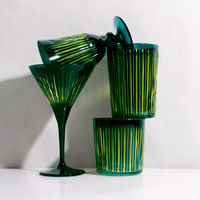 Four green-tinted, hand-cut Prism DOF glasses by L'Objet, featuring vertical grooves, are stacked artistically against a light background. One is a martini glass; the other three are tumblers—two stacked and one leaning on top.
