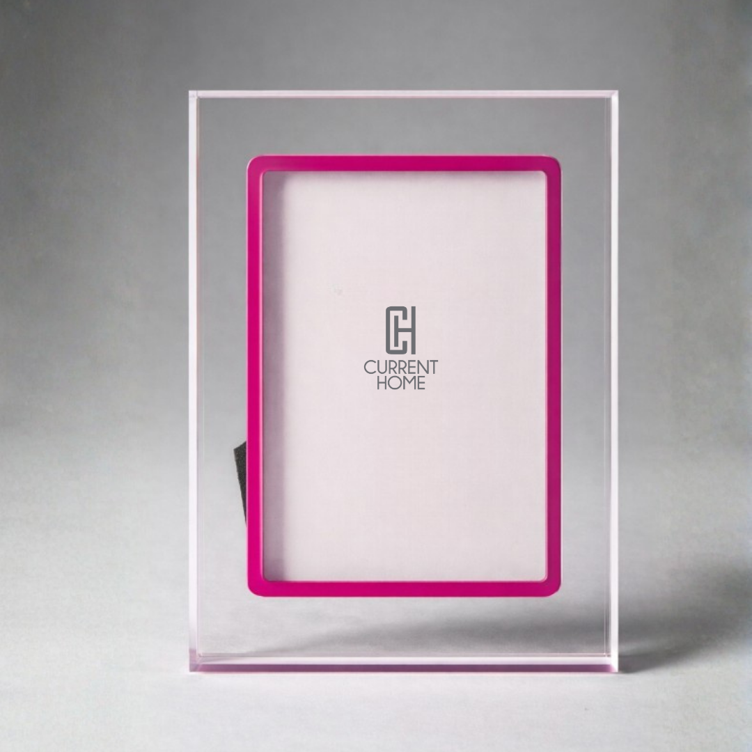 The Prisma Lip Frame - Fuschia by Bella Moulding is a clear acrylic picture frame with a bright pink inner border, shown upright on a light gray surface and displaying a placeholder with the Current Home logo in the center.