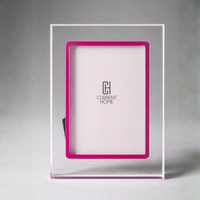 The Prisma Lip Frame - Fuschia by Bella Moulding is a clear acrylic picture frame with a bright pink inner border, shown upright on a light gray surface and displaying a placeholder with the Current Home logo in the center.