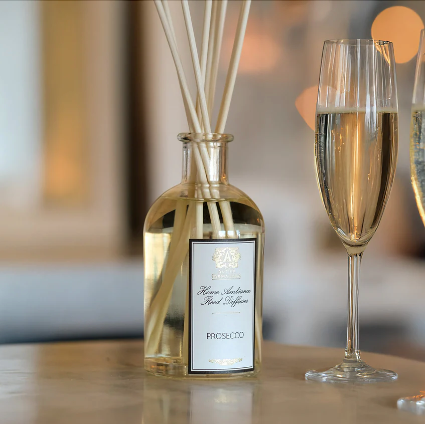 A glass of sparkling prosecco sits beside the Antica Farmacista Prosecco Diffuser by Harper on a marble surface, filling the air with an elegant home fragrance against a softly blurred background.