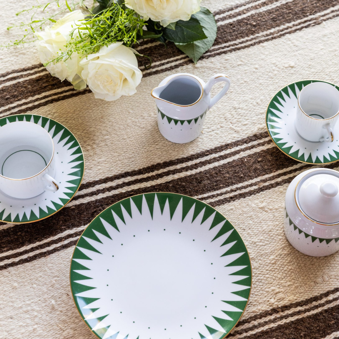 The Molecot Punk Dinnerware Green set, featuring hand-painted porcelain with a green and white starburst design, including a plate, cups, pitcher, and sugar bowl, is displayed on a striped beige-brown tablecloth beside white roses.