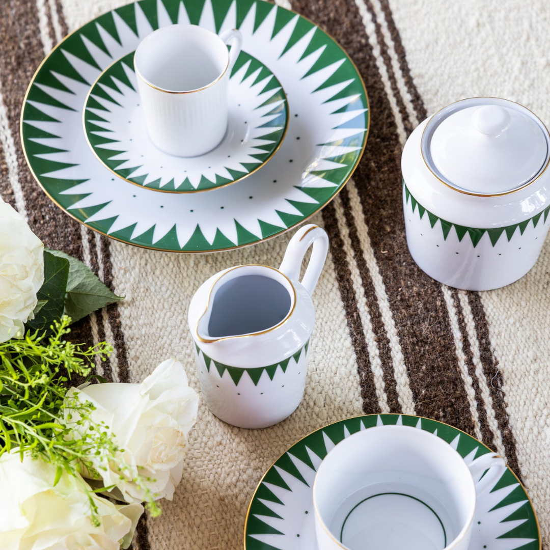 The Punk Dinnerware Green set by Molecot, featuring green and gold geometric patterns on white porcelain, is arranged on a woven brown and beige tablecloth next to a bouquet of white flowers. The set includes plates, cups, a sugar bowl, and creamer.