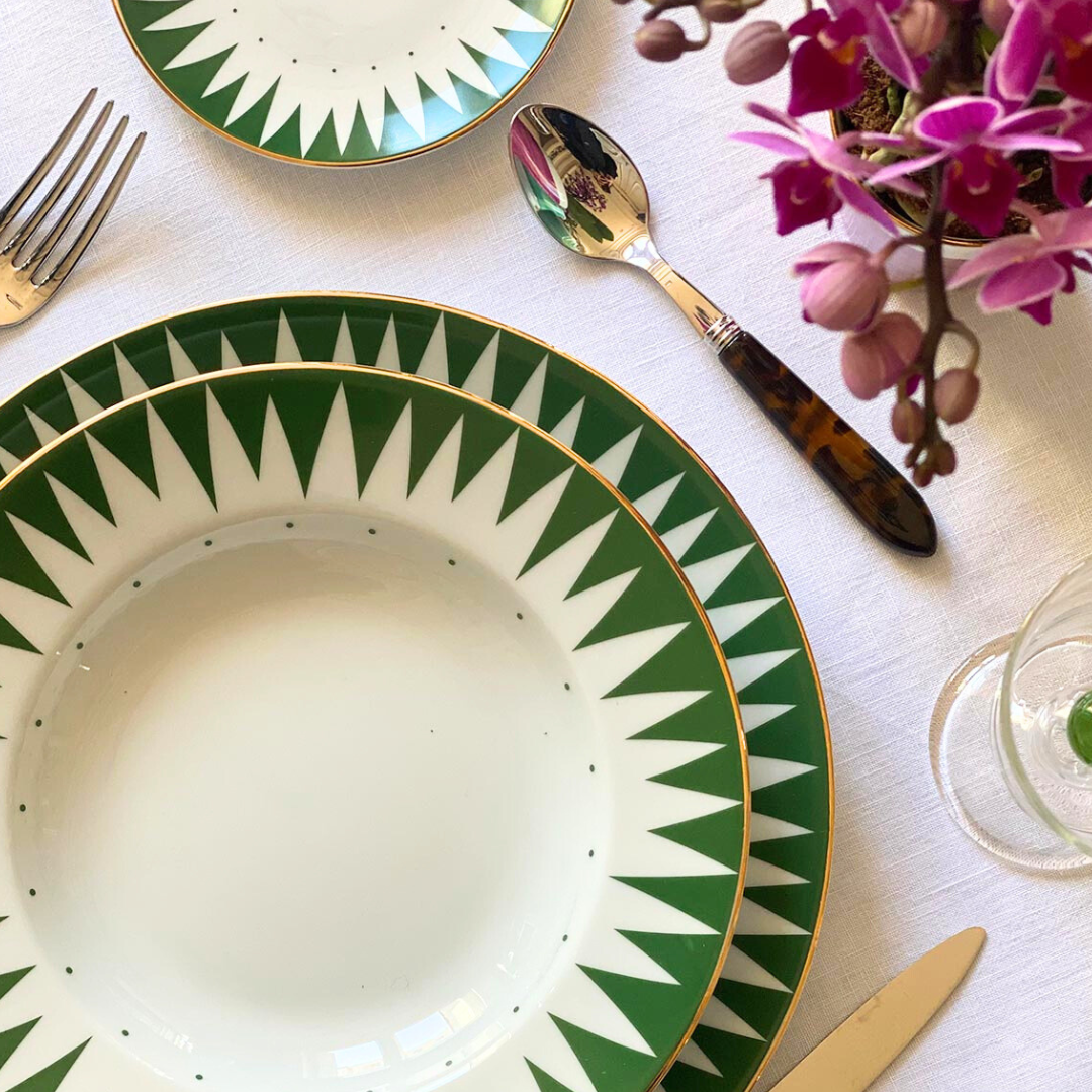 A close-up of an elegant table set with Molecot’s Punk Dinnerware Green hand-painted porcelain plates, silverware, a pink orchid arrangement, and part of a wine glass on a white tablecloth.