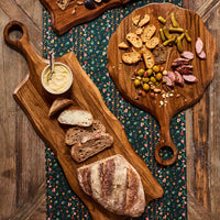 Two rustic Juliska Puro Long Serving Boards, each crafted from solid teak wood, are arranged on a wooden table adorned with a floral-patterned cloth runner—one board holds bread and butter, while the other displays sliced sausage, pickles, olives, nuts, and crackers—enhancing the natural charm of this inviting spread.