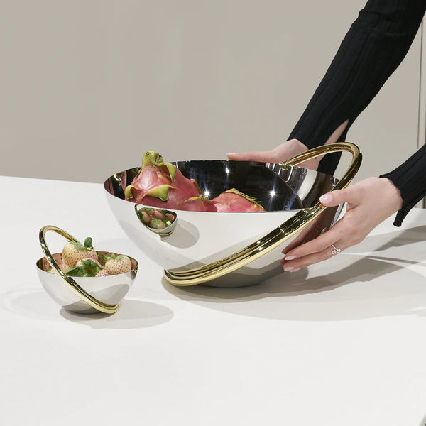 A person arranges the Anna New York Ring Fruit Bowl, a shiny stainless steel piece with gold accents filled with dragon fruit, as a centerpiece beside a matching smaller bowl holding strawberries on a white surface.