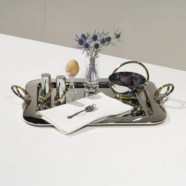 A polished stainless steel tray holds a vase with purple flowers, Anna New York Ring Salt & Pepper Shaker Set, an egg in a cup, a napkin with a spoon, and a bowl with brass handle on a white surface.