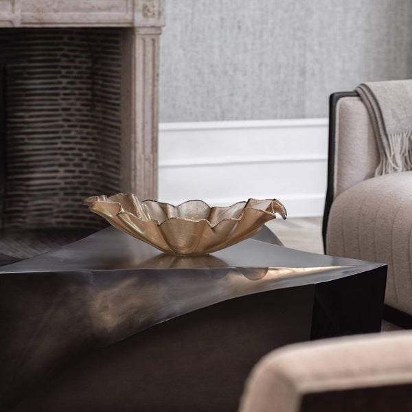 An Anenoma Bowl with copper and bronze trim by Gardeco sits on a dark, angular coffee table in a modern living room with a fireplace, a beige armchair, and a light gray wall.