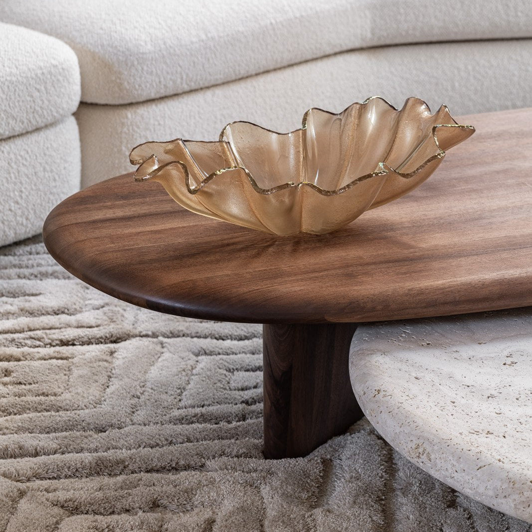 A wavy, handcrafted Anenoma Bowl with copper and bronze trim by Gardeco sits on a dark wooden coffee table. The table is placed on a textured, light-colored rug, with a curved, cream-colored sofa in the background.