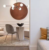 A modern dining area features a round marble table, gray chair, Lirio R Matt Black & Bronze Bowl by Gardeco, beige rug, and wall art. A white couch with mustard and striped pillows sits beneath glass art by Regina Medeiros.