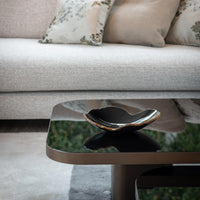 A modern black glass coffee table displays the Gardeco Mapa Bowl Black & Bronze Trim - Mini, set before a light gray sofa with floral cushions. The table’s surface beautifully reflects the decorative piece and its surroundings.