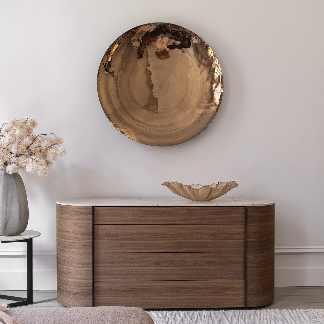 A round hammered metal wall art piece hangs above a curved handcrafted wooden sideboard with a marble top. The Anenoma Bowl - Copper & Bronze Trim by Gardeco sits on the sideboard, and a vase with pale flowers is on a small side table nearby.