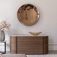 A round hammered metal wall art piece hangs above a curved handcrafted wooden sideboard with a marble top. The Anenoma Bowl - Copper & Bronze Trim by Gardeco sits on the sideboard, and a vase with pale flowers is on a small side table nearby.