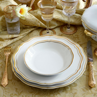 A formal place setting features Raynaud’s Polka Gold Dinnerware—white, gold-rimmed plates and a scalloped bowl—paired with gold cutlery and a water glass adorned with a flower.