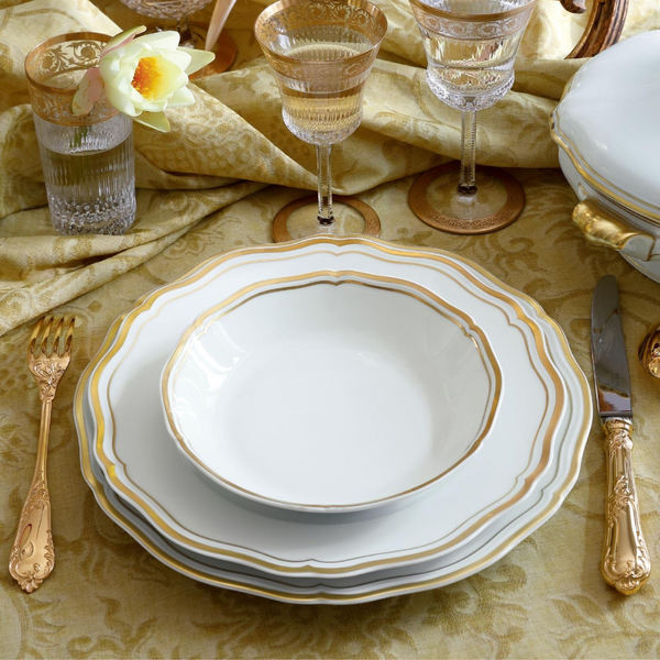 A formal place setting features Raynaud’s Polka Gold Dinnerware—white, gold-rimmed plates and a scalloped bowl—paired with gold cutlery and a water glass adorned with a flower.