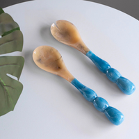 The Beatriz Ball Resin Rio Bubble Salad Servers feature beige, marbled bowls and chunky blue bubble handles, displayed on a light gray surface beside a green monstera leaf.