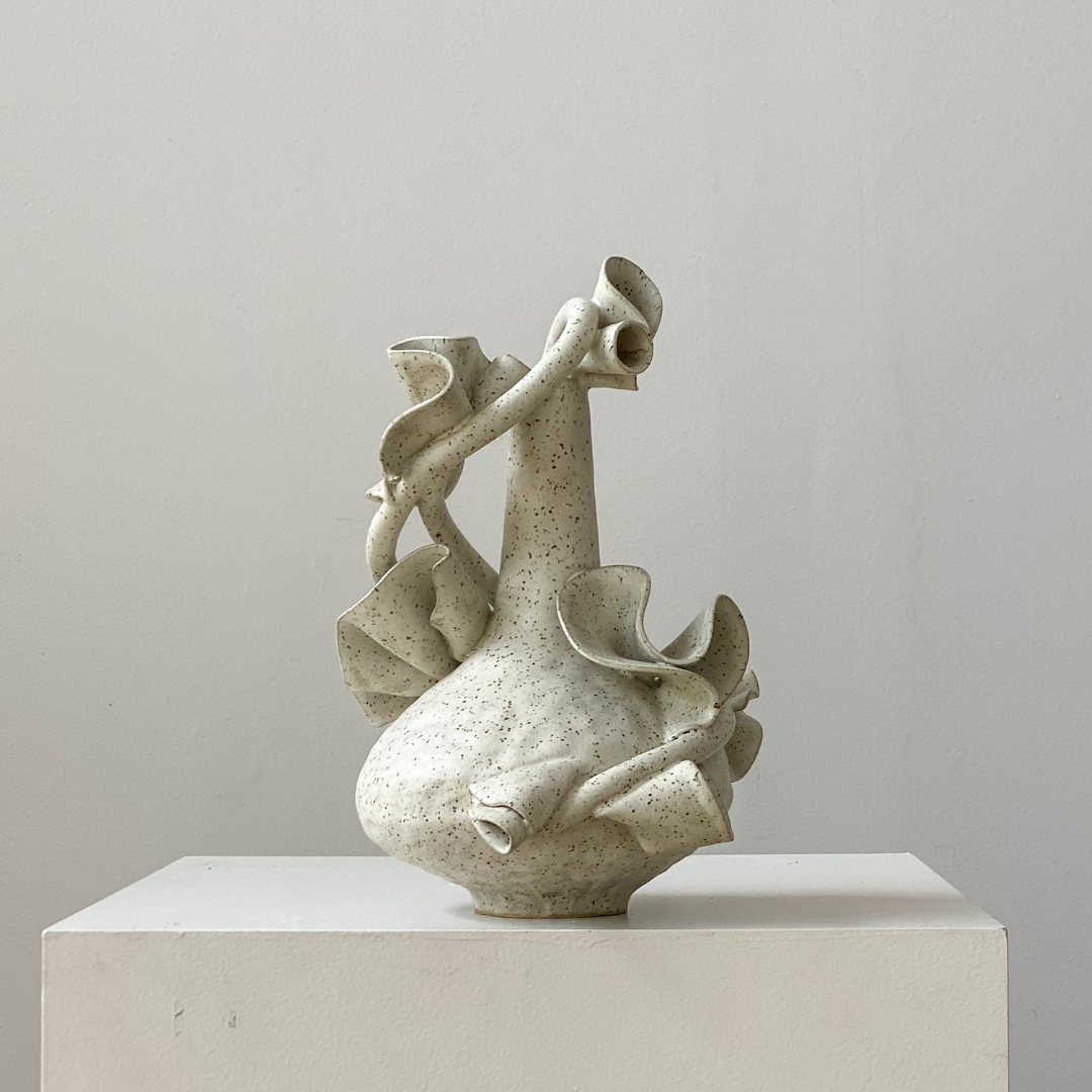The Latch Ribbon White Vessel by Latch Key Ceramics features a speckled off-white ceramic body with an organic form and ruffled edges, displayed on a white pedestal against a plain light gray background.