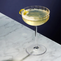 A Richard Brendon Fluted Coupe holds a pale yellow sparkling cocktail with a lemon twist, set on white marble against a dark blue background.