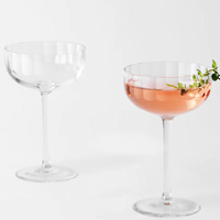 Two Richard Brendon Fluted Coupe glasses are displayed against a white background; one is empty, while the other holds a pink cocktail with a greenery garnish—both expertly crafted from premium mouth-blown glass.