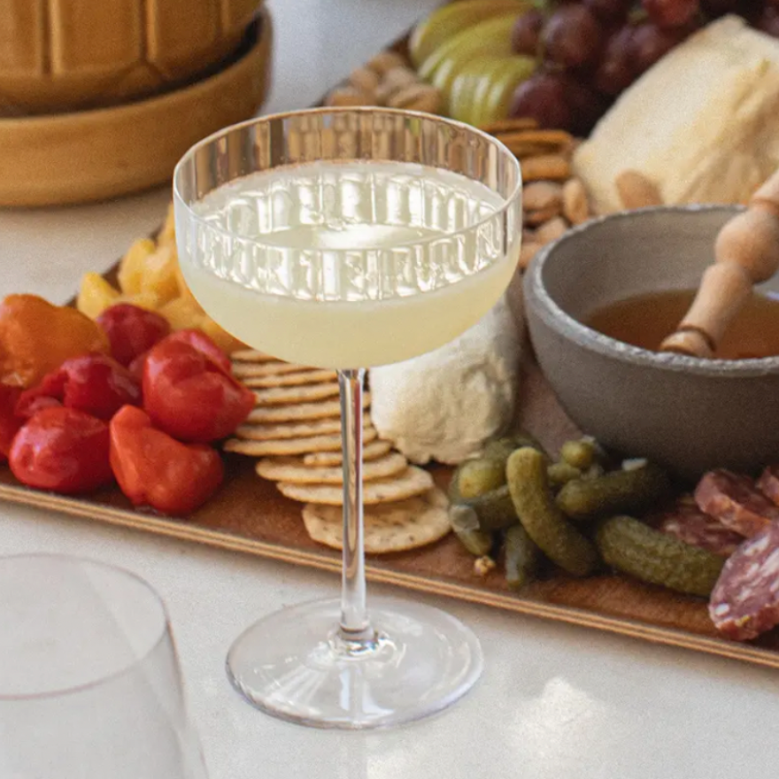 A Richard Brendon Fluted Coupe is placed in front of a charcuterie board featuring crackers, cheese, salami, cherry tomatoes, pickles, fruit, and a bowl of honey with a wooden honey dipper.
