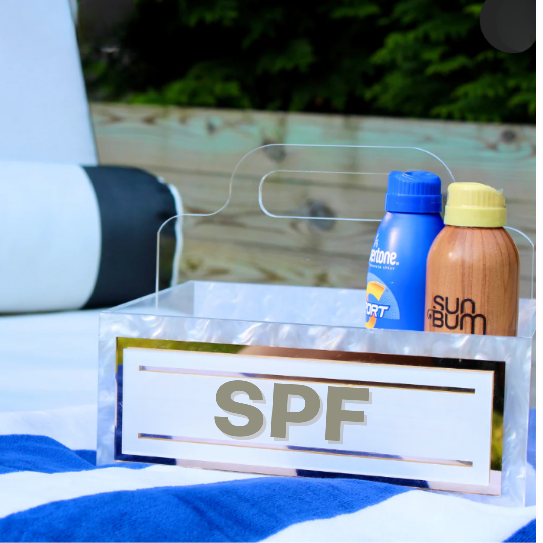 The Eye Candy Porter Acrylic Caddy - SPF holds two sunscreen bottles—one blue/yellow, one brown—set on a blue and white striped towel outdoors with greenery in the background.