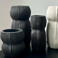 Four Curvy Round Vase Black pieces by CYM Warkov Ceramics—three in black, one in white—with wavy, ribbed surfaces sit on a black surface against a light gray background, highlighting the elegance of this artisan pottery.