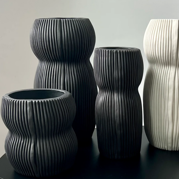 Four Curvy Round Vase Black pieces by CYM Warkov Ceramics—three in black, one in white—with wavy, ribbed surfaces sit on a black surface against a light gray background, highlighting the elegance of this artisan pottery.
