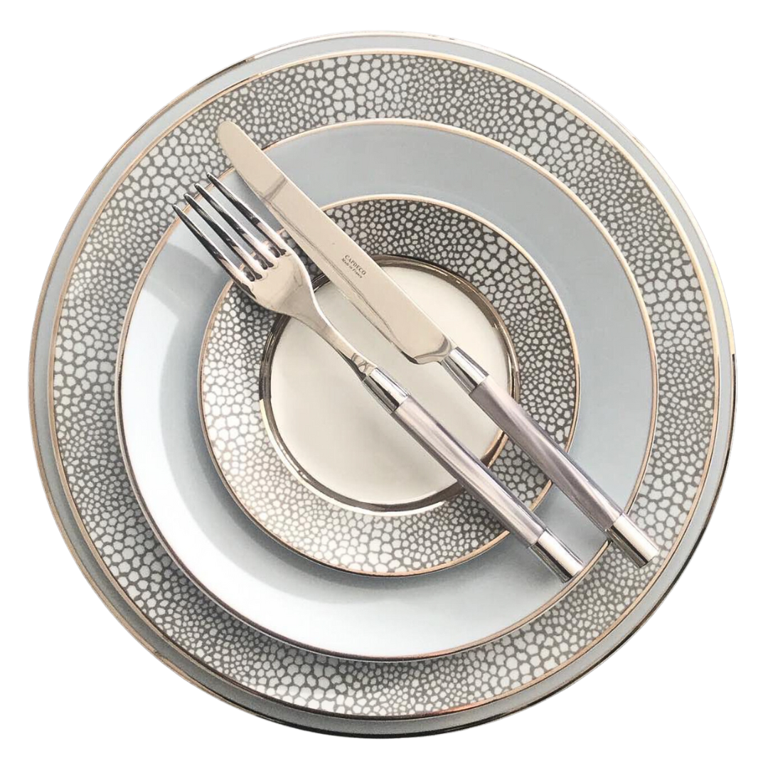 Four stacked Makassar Gray & Platinum dinner plates by Royal Limoges/Bia Cordon Bleu, crafted in France from fine Limoges porcelain with a gray and white pattern; a metallic fork and knife rest diagonally on top.