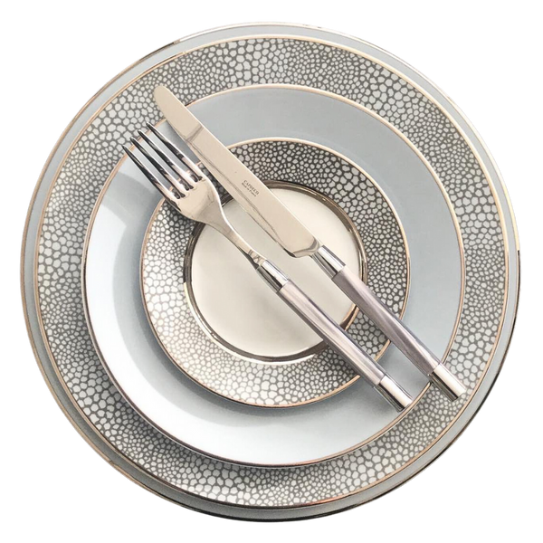 Four stacked Makassar Gray & Platinum dinner plates by Royal Limoges/Bia Cordon Bleu, crafted in France from fine Limoges porcelain with a gray and white pattern; a metallic fork and knife rest diagonally on top.
