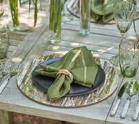 A rustic outdoor table features a black plate on the Kim Seybert Weathered Pine Placemat Ivory and Natural, with green napkin and ring, green glassware, cutlery, and tall glass vases with greenery enhancing the dining decor.