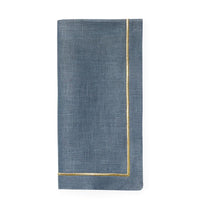 The Sferra Seaton Napkin Flint & Gold Set of 4 features neatly folded blue linen napkins with thin gold borders and pearl stitch embroidery—ideal for elegant table settings. Displayed against a white background.