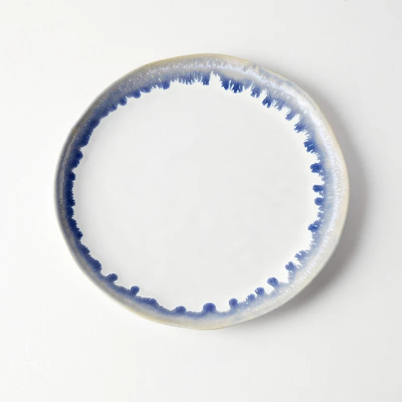 A round ceramic plate from Simon Pearce’s Burlington Dinnerware Pool collection showcases a white center accented by a blue, watercolor-inspired ring along its slightly irregular glazed edge, lending it an organic appearance when displayed on a white background.