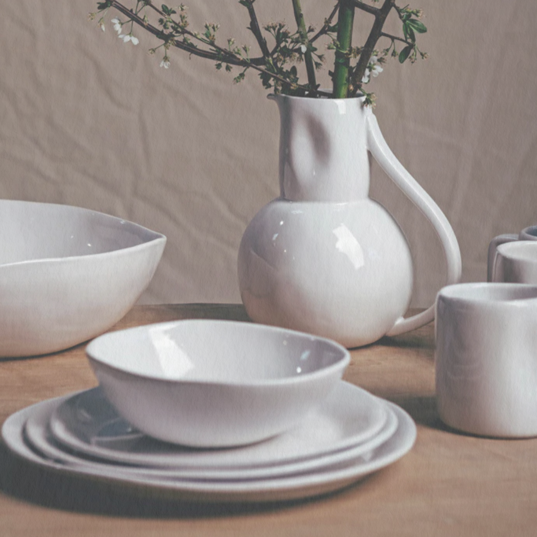 A white ceramic pitcher with branches and blossoms stands among Alex Marshall Studios' Slim Dinnerware Matte White—bowls, plates, and mugs—on a wooden table. All pieces are microwave and dishwasher safe.