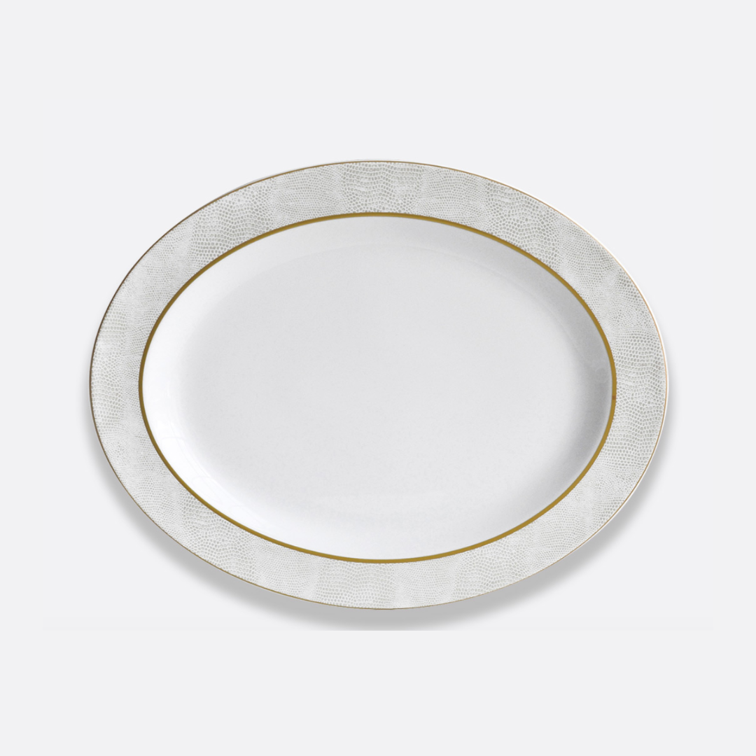 The Bernardaud Sauvage Gold Dinnerware features a white oval Limoges porcelain plate with a textured, light gray and gold snake skin-inspired rim on a plain light background.