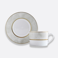 The Bernardaud Sauvage Gold Dinnerware features a white Limoges porcelain cup with handle and saucer, both adorned with light gray snakeskin-textured patterns and elegant gold trim.