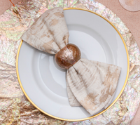 A white plate with a gold rim displays a bow-shaped beige napkin, secured by the Shell Pearl Rose Napkin Ring from Von Gern Home. The plate sits atop an iridescent, textured placemat.