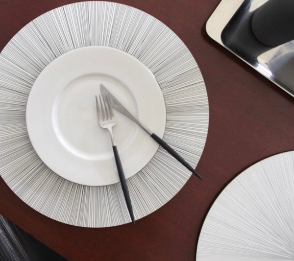 A white plate with a fork and knife rests on a round Tisch New York Lines Shadow Placemat, highlighting its bold radial line design.