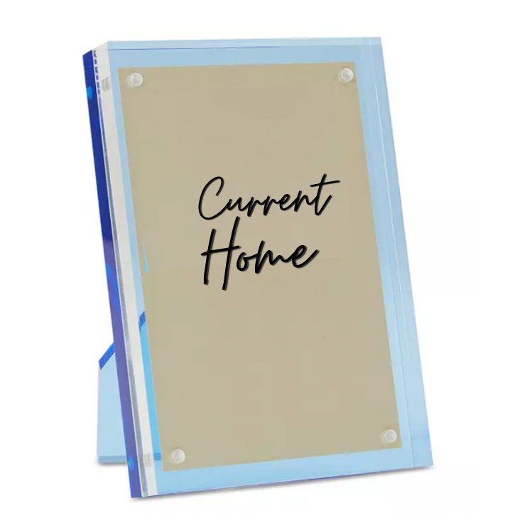 The Tizo Lucite Magnetic Frame Blue is a clear acrylic photo frame with a beige insert, displaying "Current Home" in black script. It stands upright and features sleek blue edges for a modern look.