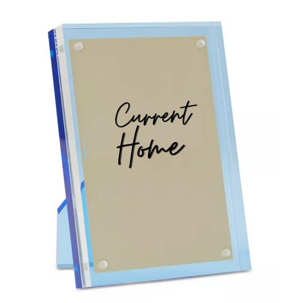 The Tizo Lucite Magnetic Frame Blue is a clear acrylic photo frame with a beige insert, displaying "Current Home" in black script. It stands upright and features sleek blue edges for a modern look.