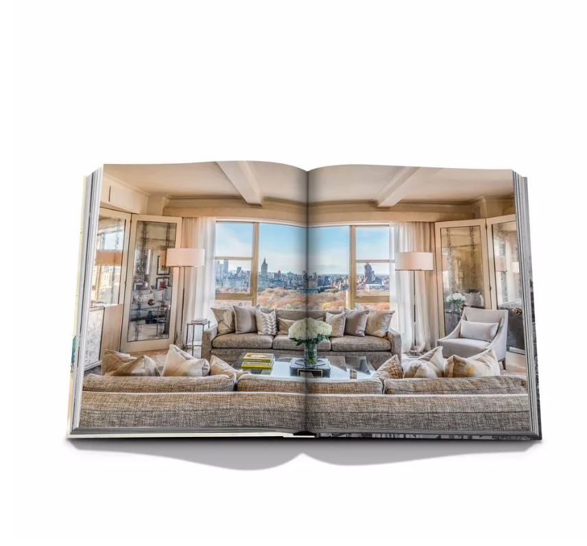 An open book, The Carlyle by Assouline, shows a modern living room with large windows overlooking a city skyline, styled with neutral sofas, a glass coffee table, and decorative lamps—evoking luxury hotel suite elegance.